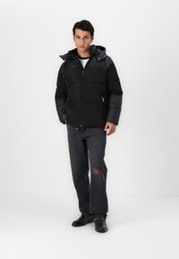 Sort puffetjakke med lynlås, aftagelig hætte og kontrasterende ærmepaneler. Sammen med grå slidte jeans og sorte støvler.