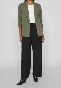 Blazer vert olive à la coupe ajustée, manches longues et deux poches avant, associé à un pantalon noir large à taille haute et des bottes noires pointues.