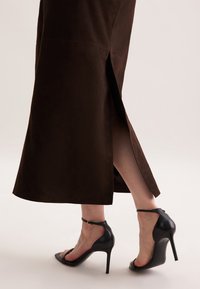 Jupe en suède brun, longueur cheville avec fente sur le côté. Une paire de sandales à talons hauts noires avec une bride de cheville et un bout ouvert visible à la base.