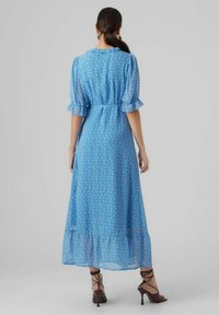 Kvinna med mörkt hår uppsatt i en ljusblå, ankellång klänning med prickar, volangärmar och bruna högklackade sandaler, stående.