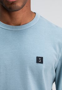Hellblaues Langarm-T-Shirt aus weichem Stoff, mit einem gerippten Rundhalsausschnitt und einem schwarzen quadratischen Patch mit einem weißen "J"-Logo.