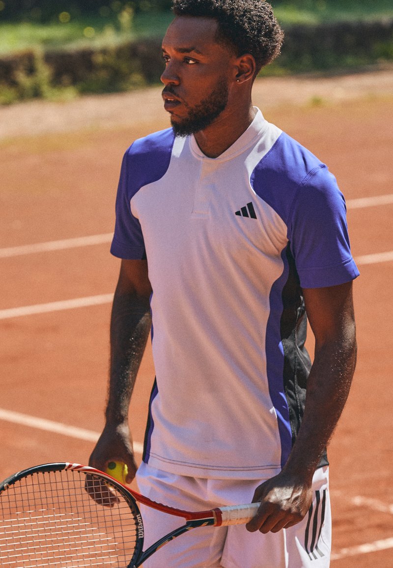 Atletisch poloshirt in het wit met paarse en zwarte details, korte mouwen en een kraag. Houdt een tennisracket vast op een gravelbaan.