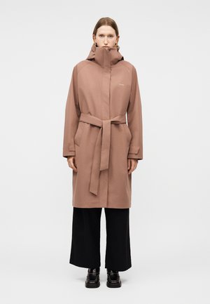 Γυναίκα που στέκεται με το σώμα στραμμένο προς τα εμπρός, φοράει καφέ παλτό με κουκούλα και ζώνη πάνω από μαύρο παντελόνι φαρδιάς γραμμής και μαύρα παπούτσια, μπροστά σε απλό φόντο.