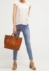 Hnědá kožená kabelka s dvojitými uchy, zvlněný světle růžový vršek, modré skinny džíny a béžové sandály na podpatku. Hladké textury a jemné detaily.