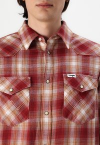 Rote und weiße karierte Flanellhemd mit Druckknöpfen, spitzem Kragen und zwei Vorderaschen; gewebtes Etikett auf der linken Tasche sichtbar.