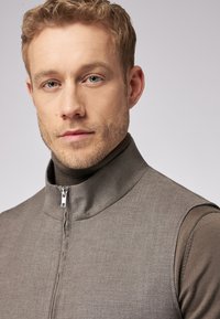 Graue Weste mit Reißverschluss und hohem Kragen aus strukturiertem Stoff, kombiniert mit einem braunen Rollkragenpullover. Die Weste zeichnet sich durch klare Linien und minimale Details aus.