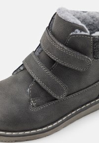 Bota sintética gris para el tobillo con dos correas de Velcro, un forro suave de vellón y costuras en contraste a lo largo de la suela. Diseño de punta redonda.