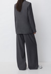 Costume à fines rayures grises avec un blazer ajusté et un pantalon à jambes larges, présentant une texture lisse et un design minimaliste. Des baskets blanches sont portées.