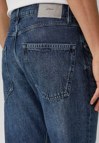 Jeans in denim di colore blu scuro, con una finitura testurizzata, tasche posteriori standard e una patch in pelle grigia con il marchio all'altezza della vita.