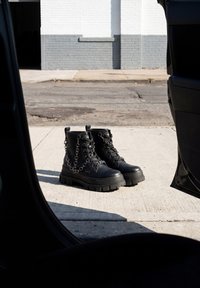 Paire de bottes de combat en cuir noir avec des chaînes posées sur le trottoir, vues à travers les portières ouvertes d'une voiture dans une rue urbaine.