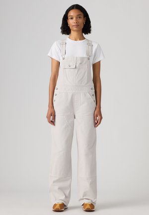 Jeune femme aux cheveux bouclés portant un t-shirt blanc, une salopette beige clair et des baskets marron, debout devant un fond uni.