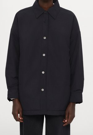 Veste noire à manches longues avec boutons et col portée par une personne, vue de face, mains détendues sur les côtés, fond uni.
