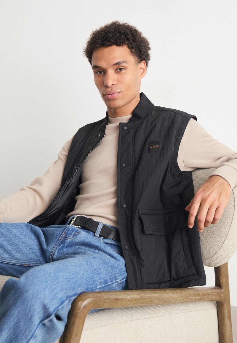 Ein junger Mann sitzt auf einem beigen Stuhl, trägt eine schwarze Weste über einem beigen Pullover und blauen Jeans und schaut ruhig in die Kamera.