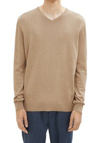 Uomo che indossa un maglione beige a maniche lunghe con scollo a V, con polsini e orlo a coste, abbinato a pantaloni blu scuro, su uno sfondo bianco.