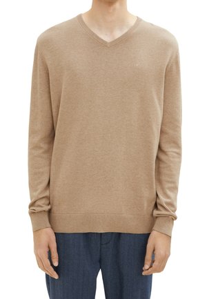 Mies, joka käyttää vaaleaa beigeä V-kaula-aukkopaidasta, jossa on ribbireunat hihoissa ja helman ympärillä, yhdistettynä tummansinisiin housuihin, valkoista taustaa vasten.