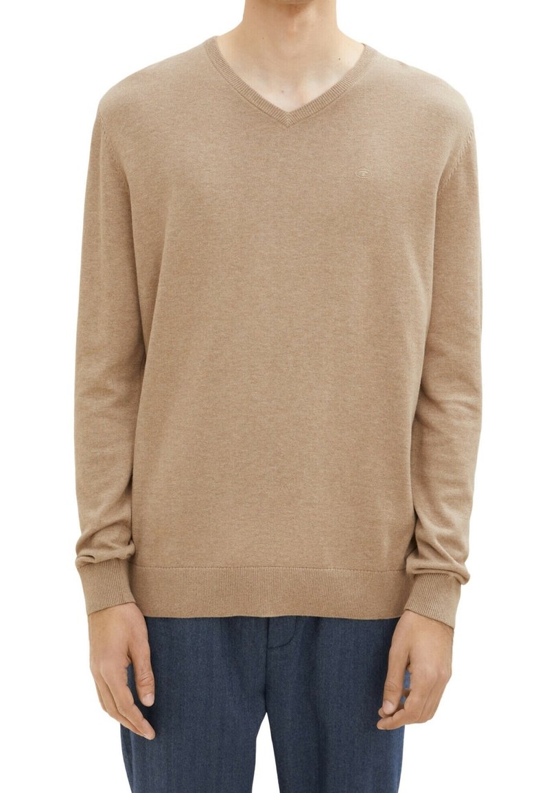 Uomo che indossa un maglione beige a maniche lunghe con scollo a V, con polsini e orlo a coste, abbinato a pantaloni blu scuro, su uno sfondo bianco.