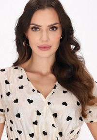 Blusa color crema con motivi a cuore neri, tessuto leggero e strutturato, scollatura a V e maniche corte arricciate.