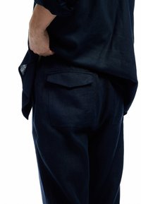 Marineblaue Leinenhemd mit lockerem Schnitt, ausgestattet mit einem abgerundeten Saum und einer detaillierten Rückentasche. Die glatte Textur hat einen leichten Schimmer.