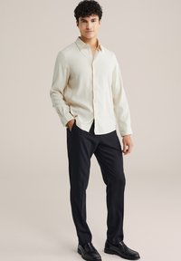 Svetlo béžová košeľa s dlhým rukávom a zapínaním na gombíky, spárovaná s čiernymi nohavicami. Košeľa má hladkú textúru a klasický golier.