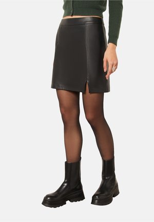 Jupe mini en cuir noir avec une silhouette ajustée, présentant un design à fente à l'avant. Modèle associé à des bottines noires à semelle épaisse.