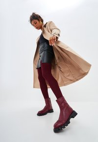 Bottines en cuir bourgogne avec des semelles noires épaisses, portées avec une jupe en cuir noire ajustée, des collants bordeaux et un trench beige.