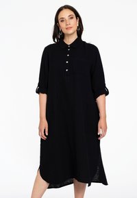 Schwarzes, strukturiertes Kleid mit kragenähnlichem Ausschnitt, Knopfleiste und Dreiviertelärmeln. Enthält eine Brusttasche und Seitenschlitze.