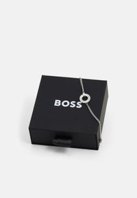 Bracciale d'argento con ciondolo rotondo, dotato di un accento nero e dettagli in cristallo, esposto su una scatola nera etichettata 'BOSS.'