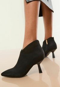 Botines de ante negro con punta afilada, tacón stiletto de altura media y cierre de cremallera en la parte trasera. Textura suave y diseño elegante.