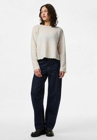Femme aux cheveux bouclés portant un pull beige, un jean large foncé et des bottes noires, debout les mains détendues.