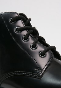 Stivale in pelle nera con lacci neri rotondi, occhielli in metallo e una texture liscia e lucida. Primo piano sulla zona dei lacci e sui dettagli delle cuciture.