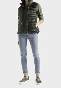 Vadderad svart väst med hög krage, grön skjorta under, ljusblå jeans och gröna sneakers med vita sulor. Dragkedja och logodetalj synlig.