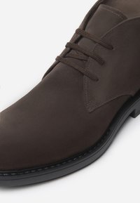Stivaletti alla caviglia in suede marrone con una texture liscia, punta tondeggiante e design con lacci. Presentano una suola sintetica nera e dettagli di cucitura minimi.