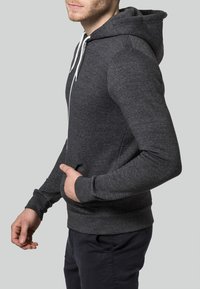 Man som bär en mörkgrå zip-up hoodie med vita snörningar och mörka byxor, står på sidan med en hand i huvan.