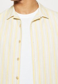 Camicetta a maniche corte con colletto a righe verticali bianche e gialle, dotata di chiusura a bottoni e texture morbida. Indossata sopra una t-shirt bianca.