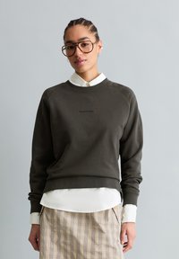 Frau mit geflochtenem Haar und Brille, die einen dunklen Pullover über einem weißen Hemd und einem beigen karierten Rock trägt, stehend vor einem grauen Hintergrund.