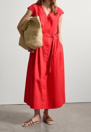 Femme portant une robe midi rouge avec une taille nouée, tenant un grand sac beige tissé, et portant des sandales plates marron, debout sur un sol en béton.