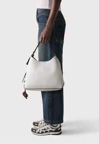 Sac à épaule en cuir blanc, de forme courbée, accentué par une sangle foncée et des pompons décoratifs. Porté avec un jean bleu et des baskets tendance.