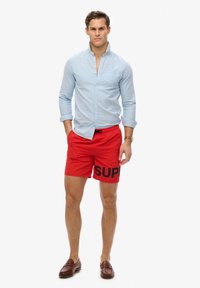 Camisa azul claro de botones combinada con pantalones de baño rojos que presentan un estampado negro de "SUP". Mocasines marrones completan el conjunto.