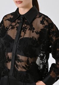 Zwarte doorzichtige blouse met bloemenborduurwerk, knopen aan de voorkant en een gestructureerde kraag. Bevat textuur met bloemmotieven en een fitted taille.