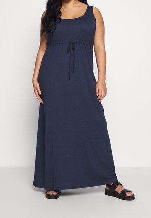 Frau trägt ein marineblaues ärmelloses Maxikleid mit Kordelzug an der Taille und schwarzen Plateausandalen vor einem einfarbigen weißen Hintergrund.