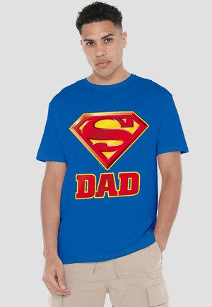 Young man wearing blue T-shirt with red and yellow Superman logo above the word "DAD," paired with beige cargo shorts.