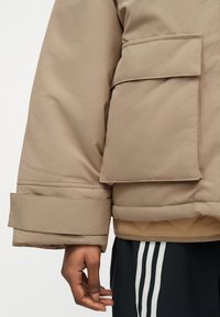 Κοντινό πλάνο ενός ατόμου που φοράει μπεζ μπουφάν με επένδυση και μεγάλη τσέπη με καπάκι, καθώς και μαύρο παντελόνι με λευκές ρίγες.