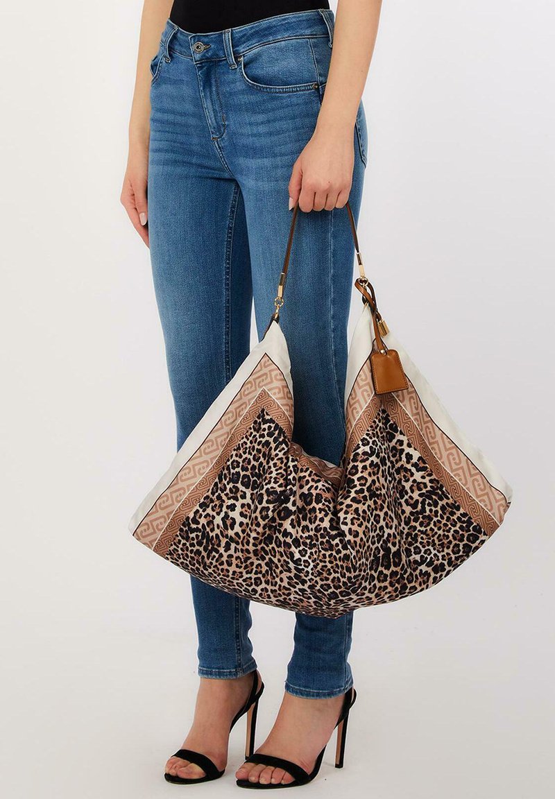 Mujer con vaqueros azules y tacones negros sostiene un gran bolso tote con estampado de leopardo y borde marrón por sus asas marrones.