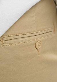Beige bomullsbyxor med en platt bakficka med en sydd kant och en ljus knapp. Vit skjorttyg delvis synligt ovanför.