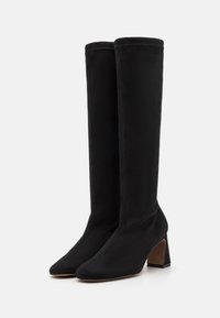 Sorte suede støvler til knæene med spidse tæer, chunky hæle og syninger langs sømmene. Designet til en tætsiddende silhuet.