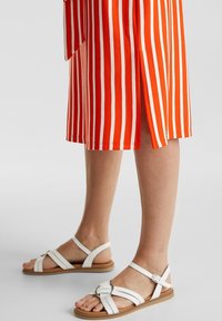 Białe płaskie sandały z wieloma paskami, w tym szerokim paskiem związanym z przodu i z zapięciem na klamrę. Noszone z czerwono-białą paskowaną spódnicą.