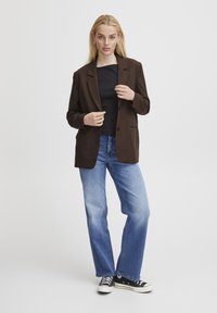 Blazer brun ajusté avec revers en pointe, fermeture à un bouton, et deux poches avant, associé à un jean bleu à jambes larges et des baskets noires.
