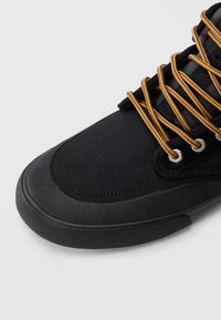 Černý teniska s texturovanou gumovou podrážkou a žluto-hnědými tkaničkami protaženými stříbrnými očky na bílém pozadí.