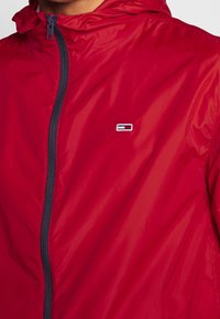 Veste légère rouge avec fermeture éclair, capuche et une fermeture éclair contrastante bleu marine. Présente un petit logo sur la poitrine. Texture lisse.