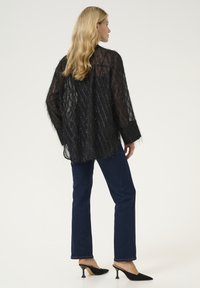 Caurspīdīga melna blūze ar fakturētu rakstu, garām piedurknēm un franjām, apvienota ar tumšām džinsām un melniem norādītām papēžiem.